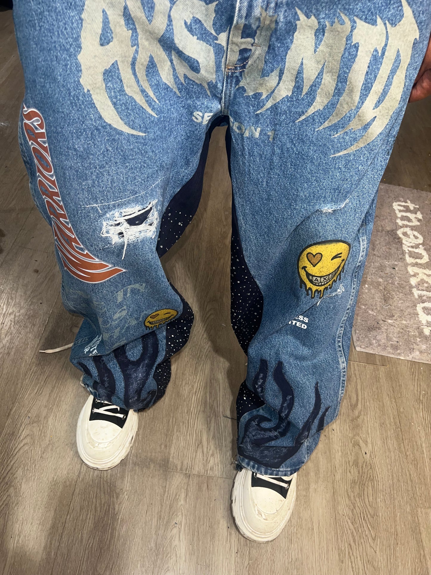 Blue jeans with graphic designs and patches worn by a person on a wooden floor.